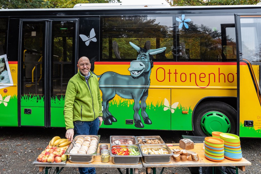 Mann in grüner Jacke steht vor einem Bus mit Esel-Illustration und einem Tisch mit Essen und bunten Tellern