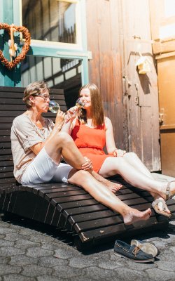 Zwei Frauen sitzen auf einer Holzliege vor einem Haus und trinken Wein aus Gläsern