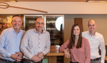 Vier lächelnde Personen lehnen an einer Bar in einem gemütlichen Raum mit Weinregal im Hintergrund.