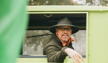 Mann mit Hut und Brille lehnt aus dem Fenster eines hellgrünen Fahrzeugs, unscharfer grüner Vordergrund