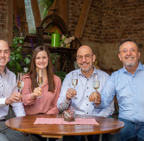 Vier Personen sitzen an einem Holztisch und stoßen mit Sektgläsern in einem rustikalen Raum an.