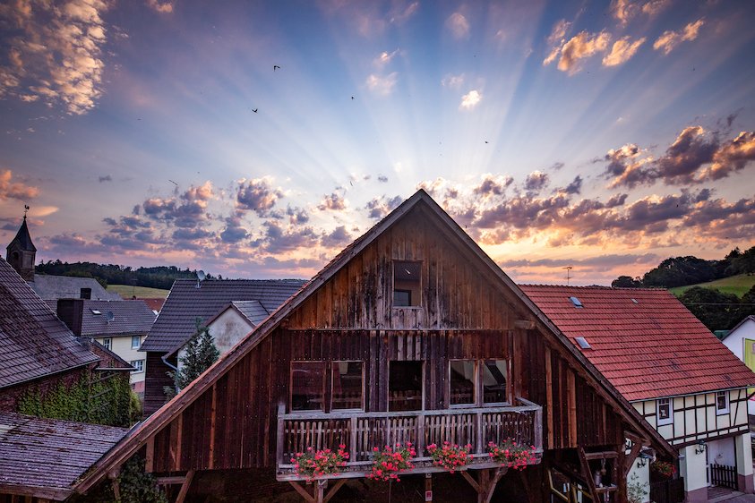 Holzhaus mit Balkon und Blumen vor Sonnenuntergang mit strahlenförmigen Wolken am Himmel