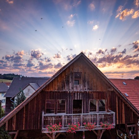 Holzhaus mit Balkon und Blumen vor Sonnenuntergang mit strahlenförmigen Wolken am Himmel