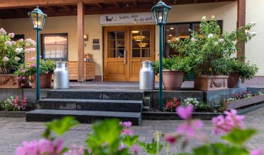 Eingang eines Hauses mit Holztreppe, zwei Milchkanistern, Blumen und Laternen