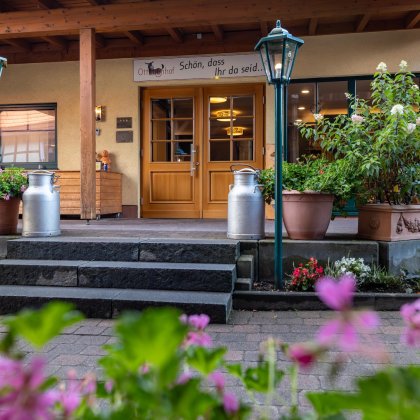 Eingang eines Hauses mit Holztreppe, zwei Milchkanistern, Blumen und Laternen