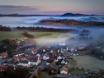 Luftaufnahme eines Dorfes mit Nebel über Feldern und bewaldeten Hügeln im Hintergrund bei Dämmerung