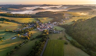 Luftaufnahme eines Dorfes in einem Tal mit Feldern und bewaldeten Hügeln im Morgennebel