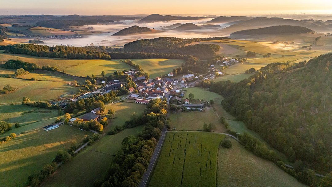Luftaufnahme eines Dorfes in einem Tal mit Feldern und bewaldeten Hügeln im Morgennebel