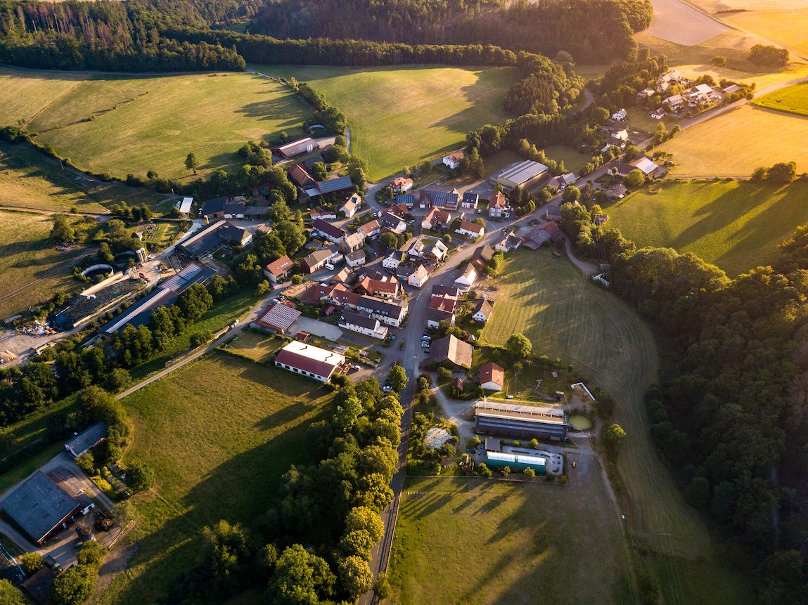 Luftaufnahme eines kleinen Dorfes mit Häusern, Feldern und Wäldern im Abendlicht