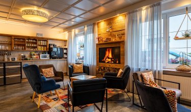 Gemütlicher Aufenthaltsraum mit Kamin, Sesseln um einen Tisch und Küchenzeile im Hintergrund