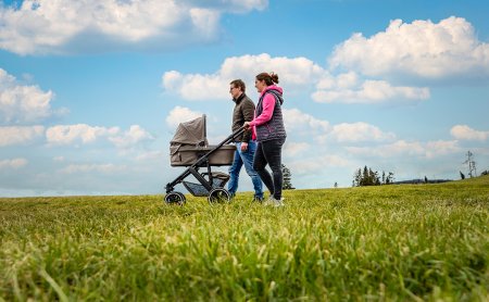 Zwei Personen schieben einen Kinderwagen über eine grüne Wiese unter blauem Himmel mit Wolken