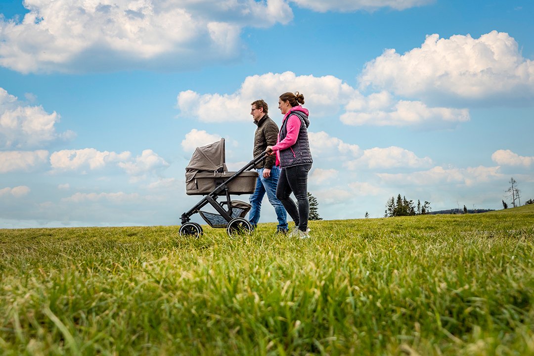 Zwei Personen schieben einen Kinderwagen über eine grüne Wiese unter blauem Himmel mit Wolken