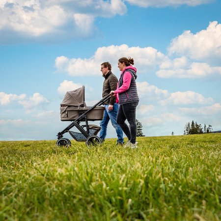 Zwei Personen schieben einen Kinderwagen über eine grüne Wiese unter blauem Himmel mit Wolken