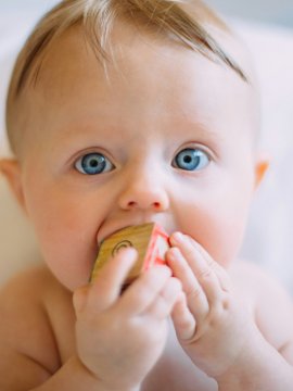 Nahaufnahme eines Babys mit blauen Augen, das einen Holzbaustein in den Mund nimmt