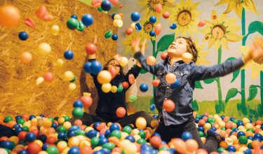 Kinder spielen in einem Bällebad vor einer Wand mit Sonnenblumenmalerei und werfen bunte Bälle in die Luft