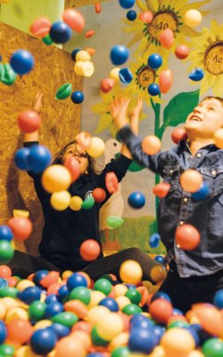 Kinder spielen in einem Bällebad vor einer Wand mit Sonnenblumenmalerei und werfen bunte Bälle in die Luft