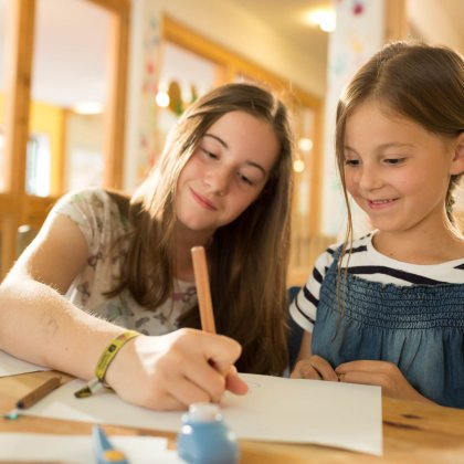 Zwei Mädchen sitzen an einem Tisch, eines zeichnet auf Papier, das andere schaut lächelnd zu