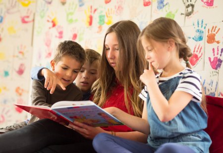 Eine Frau liest Kindern vor, die an einer Wand mit bunten Handabdrücken sitzen