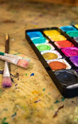 Aquarellfarben in einer Palette rechts, zwei Pinsel liegen gekreuzt auf einem bemalten Tisch links