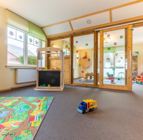 Heller Spielraum mit Teppich, Spielzeugauto und Holzmöbeln vor großen Fenstern und Glastüren