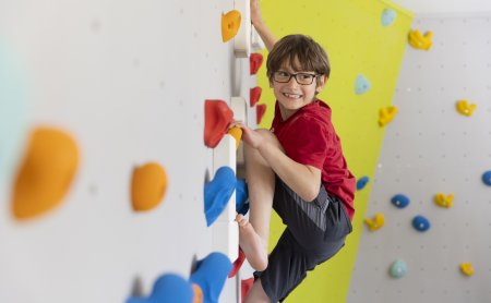 Junge mit Brille klettert an einer bunten Kletterwand und lächelt nach rechts