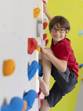 Junge mit Brille klettert an einer bunten Kletterwand und lächelt nach rechts