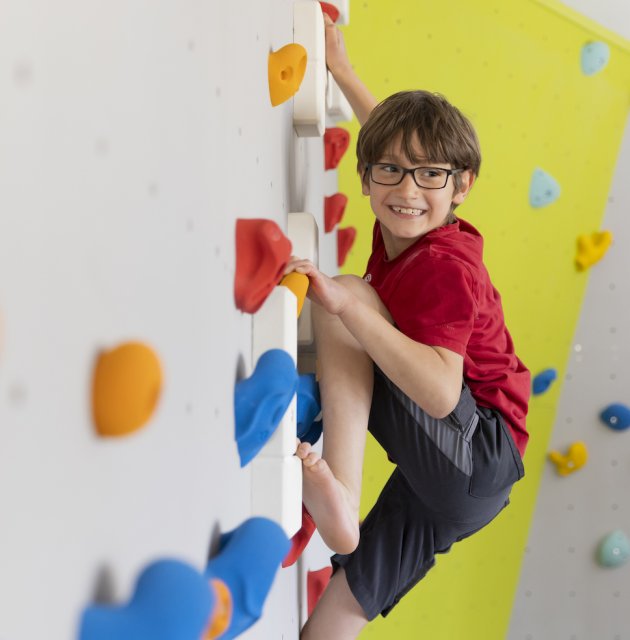 Junge mit Brille klettert an einer bunten Kletterwand und lächelt nach rechts