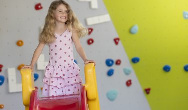 Lächelndes Mädchen steht oben auf einer roten Rutsche mit gelben Griffen vor einer Kletterwand