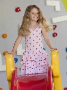 Lächelndes Mädchen steht oben auf einer roten Rutsche mit gelben Griffen vor einer Kletterwand