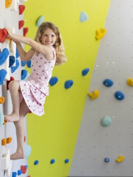 Mädchen klettert lächelnd an bunter Kletterwand mit roten, blauen und orangen Griffen