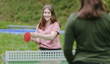 Junges Mädchen in rosa T-Shirt spielt Tischtennis draußen und hält Schläger und Ball