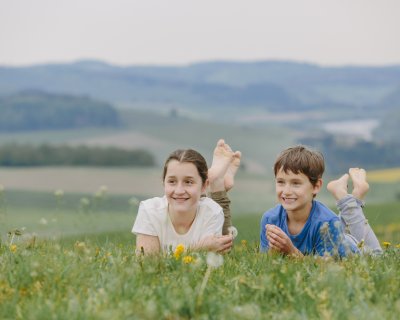 Zwei Kinder liegen auf einer Wiese mit Blumen, im Hintergrund Hügel und ein Fluss