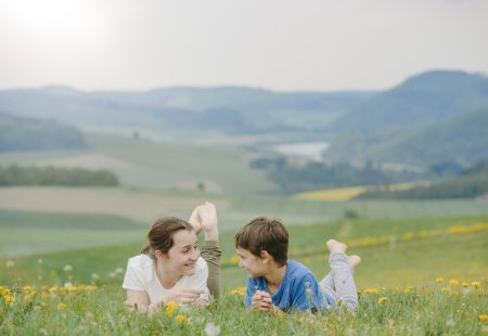 Zwei Kinder liegen auf einer Wiese mit gelben Blumen und schauen sich lächelnd an, im Hintergrund Hügel