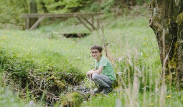 Junge mit Brille hockt rechts neben einem kleinen Bach in einer grünen Wiese mit Bäumen im Hintergrund