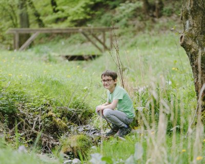 Junge mit Brille hockt rechts neben einem kleinen Bach in einer grünen Wiese mit Bäumen im Hintergrund
