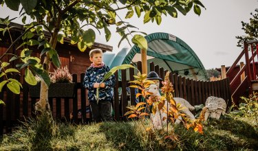 Junge steht im Garten neben Baum und Zaun, hält große Blätter, im Hintergrund Spielplatz und Gartendekoration