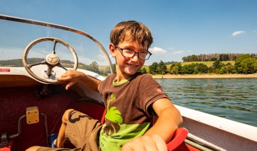 Junge mit Brille sitzt lächelnd in einem Boot am Steuer bei sonnigem Wetter auf einem See