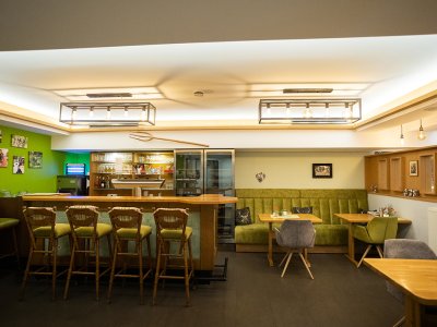 Innenraum einer gemütlichen Bar mit Holztheke, grünen Stühlen und Tischen, Wandbildern und moderner Beleuchtung