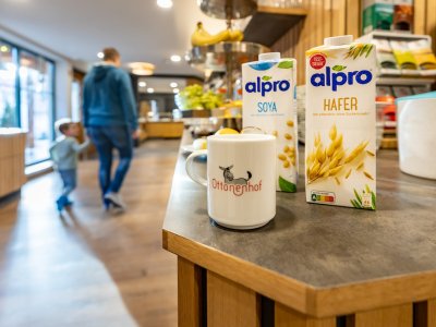 Kaffeetasse mit Ottonenhof-Logo neben Alpro Soja- und Hafermilch auf Theke, unscharfe Personen im Hintergrund