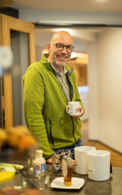 Lächelnder Mann mit Brille und grüner Jacke hält Tasse in moderner Küche