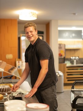 Junger Mann in schwarzem Poloshirt steht lächelnd an einem Buffet mit Kuchen und Tellern