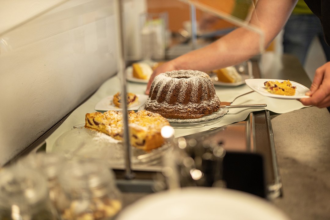 Hand nimmt ein Stück Kuchen von einer Theke mit verschiedenen Kuchen unter einer Glasabdeckung