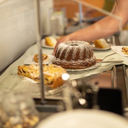 Hand nimmt ein Stück Kuchen von einer Theke mit verschiedenen Kuchen unter einer Glasabdeckung