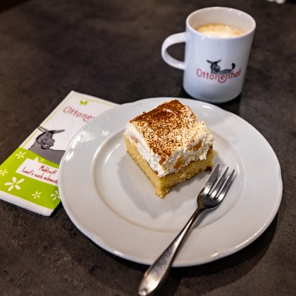 Stück Kuchen mit Sahne auf weißem Teller mit Gabel, Kaffeetasse und Serviette mit Ottonenhof-Logo