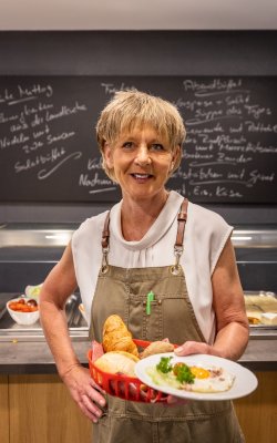 Frau in Schürze hält Teller mit Spiegelei und Korb mit Brötchen vor Theke mit Speisekarte an Wand