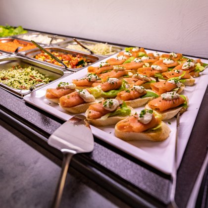 Buffet mit Lachsbrötchen auf einem weißen Teller und verschiedenen Salaten in Metallbehältern