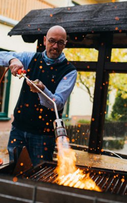 Mann mit Brille hält Gasbrenner und entzündet Feuer auf Grill im Freien