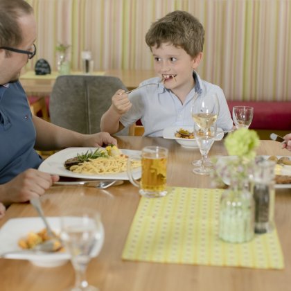 Familie sitzt an einem Tisch und isst gemeinsam, Junge isst mit Gabel und lächelt