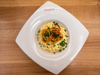 Von oben fotografierte cremige Käsespätzle mit Röstzwiebeln und Petersilie auf weißem Teller mit Aufschrift Ottoneghof