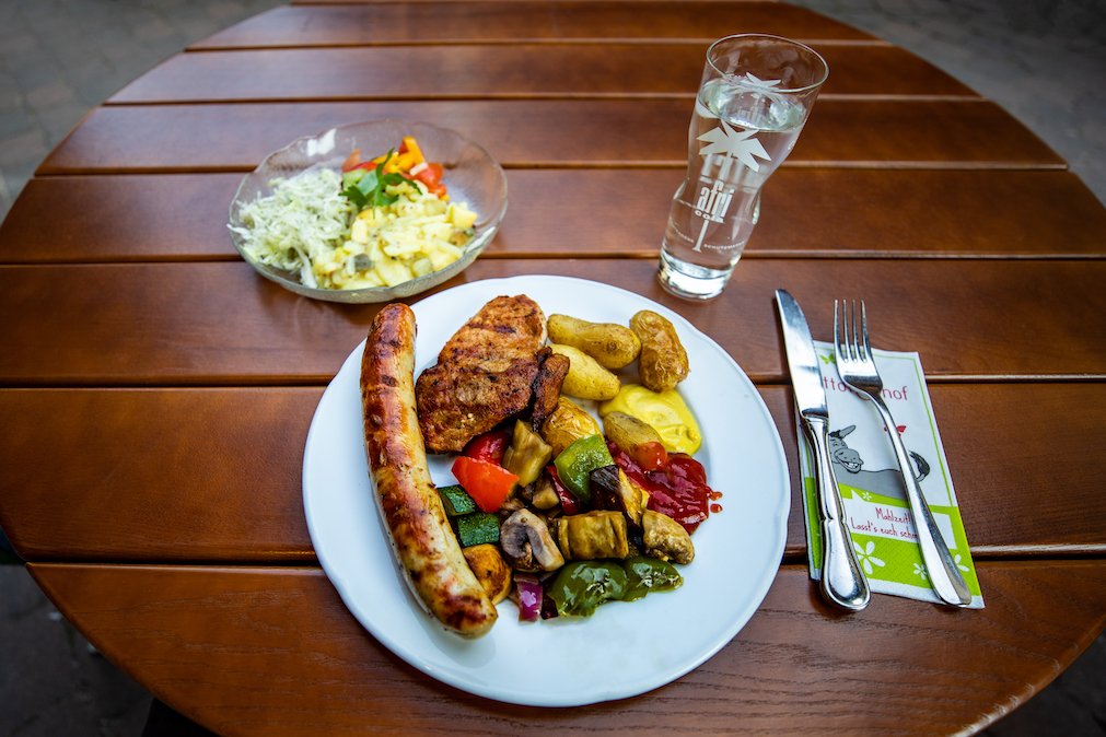 Teller mit Bratwurst, Fleisch, Gemüse, Kartoffeln und Soßen auf Holztisch, Salat und Glas Wasser daneben